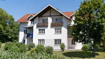 Mehrfamilienhaus mit weißer Fassade, roten Ziegeldächern und mehreren Balkonen, umgeben von viel Grün.