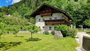 Traditionelles Haus im alpinen Stil mit großem grünen Garten und bewaldetem Berghang im Hintergrund.