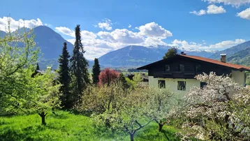 Idyllischer Blick auf das Haus mit blühenden Obstbäumen und majestätischer Bergkulisse unter blauem Himmel.