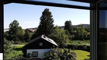 Blick aus dem Fenster auf ein Haus und die grüne, bewaldete Landschaft unter blauem Himmel.