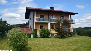 Gelbes Wohnhaus mit Balkonen und Satteldach, umgeben von grüner Landschaft unter blauem Himmel.