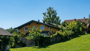 Gelbes Einfamilienhaus mit grünem Dach, umgeben von viel Grün und einem gepflegten Rasen unter blauem Himmel.