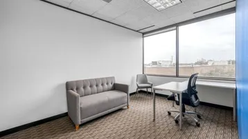 Kleines Büro mit einem Sofa, Schreibtisch und großen Fenstern, die viel Tageslicht hereinlassen.