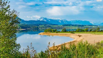 Malerischer Blick auf einen See mit schneebedeckten Bergen im Hintergrund und grüner Landschaft unter blauem Himmel.
