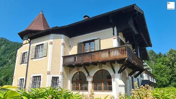 Traditionelles Gebäude mit markantem Turmdach, Holzbalkon und dekorativer Fassade unter blauem Himmel.