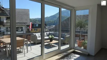 Heller Wohnbereich mit großen Glasfronten und direktem Zugang zu einer möblierten Terrasse mit Bergblick.