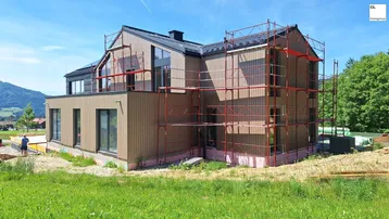 Neubau-Einfamilienhaus in Holzbauweise mit Gerüst im Bauzustand, umgeben von grüner Landschaft.
