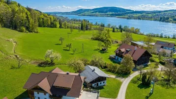 Panoramablick auf mehrere Häuser am Seeufer, umgeben von grünen Wiesen und bewaldeten Hügeln.