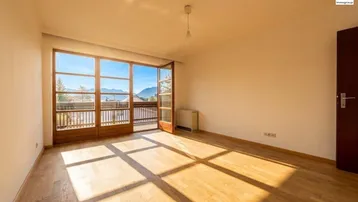 Helles Zimmer mit Holzboden und Zugang zu einem Balkon mit Bergblick, durchflutet von Sonnenlicht.
