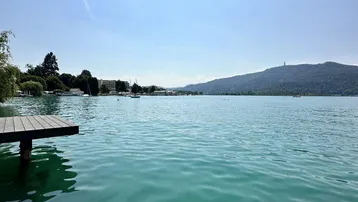 Seeufer mit Holzsteg und malerischem Blick auf die umliegende Berglandschaft unter blauem Himmel.