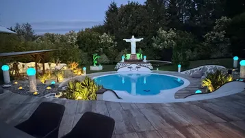 Beleuchteter Poolbereich im Garten bei Nacht mit Holzterrasse und dekorativer Statue.