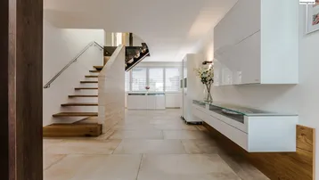 Moderner Eingangsbereich mit heller Steinfliesenboden, offener Holztreppe und schwebenden Möbeln, durchflutet von natürlichem Licht.