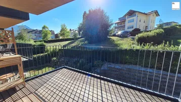 Heller Balkon mit Holzboden und Blick auf grüne Hügel und umliegende Gebäude.