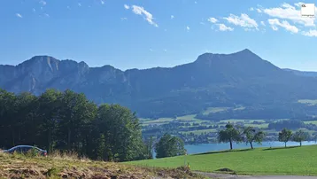 Panoramablick auf majestätische Berge, einen ruhigen See und grüne Hügellandschaft unter blauem Himmel.