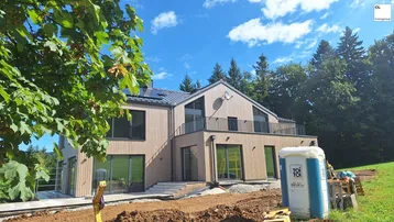 Modernes Einfamilienhaus im Bau mit Holzfassade und Blick auf die grüne Landschaft.