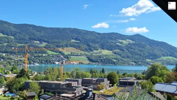 Panoramablick auf den See und die umliegenden Berge mit einigen Gebäuden im Vordergrund unter blauem Himmel.