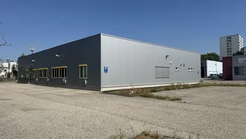 Graues Gewerbegebäude mit großem Parkplatz unter blauem Himmel und sonnigem Wetter.