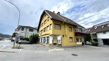 Gelbes Mehrfamilienhaus an einer Straßenecke mit Parkplätzen und bewölktem Himmel.