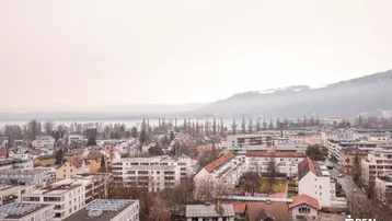 Panoramablick über die Stadt mit See und Bergen im Hintergrund an einem bewölkten Tag.