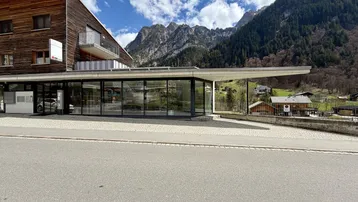 Modernes Gebäude mit Holzfassade und großen Glasfronten, eingebettet in eine alpine Berglandschaft.
