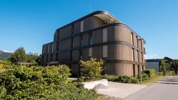 Moderne Architektur mit geschwungener Holzfassade, umgeben von gepflegter Bepflanzung.