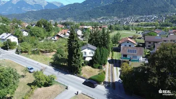 Luftaufnahme einer Wohnsiedlung mit Häusern, viel Grün und Bergen im Hintergrund.