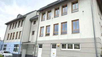 Mehrstöckiges Gebäude mit hellgrauer Fassade, vielen Fenstern und einem gepflasterten Vorplatz.
