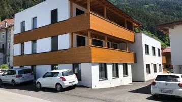 Modernes Mehrfamilienhaus mit weißer Fassade und Holzverkleidung der Balkone in bergiger Umgebung.