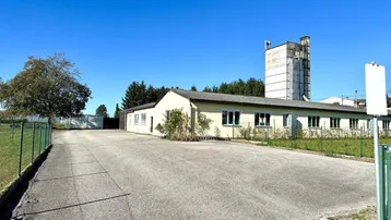 Eine weite Außenansicht des Gebäudes mit großem Parkplatz und einem hohen Silo im Hintergrund unter blauem Himmel.
