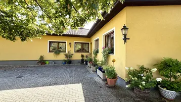 Sonnige Außenansicht des Hauses mit gelber Fassade, Fenstern und gepflastertem Hofbereich.