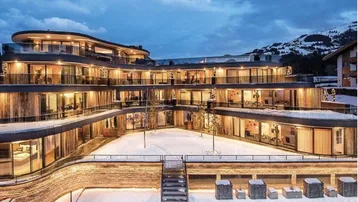 Modernes Apartmentgebäude mit mehreren Etagen, großen Glasfronten und Balkonen, umgeben von Schnee und Bergen.
