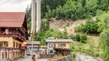 Traditionelle Gebäude am Flussufer mit einer Brücke im Hintergrund und bewaldeten Bergen.