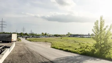 Weitläufiges Außengelände mit Zufahrtsstraße, Grünflächen und Parkplätzen im Hintergrund unter bewölktem Himmel.