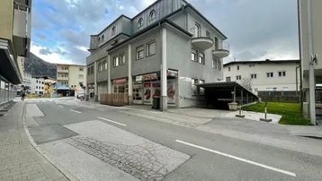Gebäudeansicht mit Geschäften im Erdgeschoss, Wohnungen darüber und einer überdachten Parkplatzzufahrt.