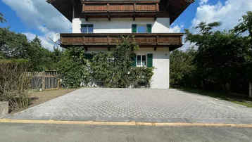 Traditionelles Haus mit Holzbalkonen und gepflasterter Einfahrt unter blauem Himmel.