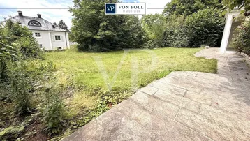 Großer Gartenbereich mit Rasenfläche und gepflasterter Terrasse, umgeben von Bäumen und einem Haus im Hintergrund.