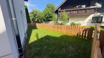 Kleiner Gartenbereich mit Rasenfläche und Holzzaun neben dem Haus, umgeben von Grün.