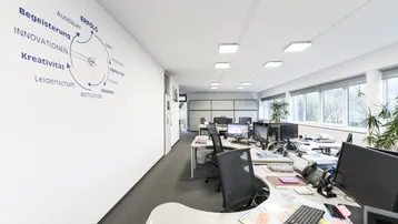 Geräumiges Großraumbüro mit mehreren Arbeitsplätzen, Teppichboden und großen Fenstern für viel Tageslicht.