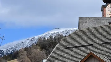 Panoramablick auf schneebedeckte Berge und traditionelle Dächer mit Kirchturm in der Ferne.