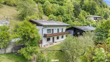 Traditionelles Haus mit Balkon und Satteldach, umgeben von grünen Hügeln und Bäumen.