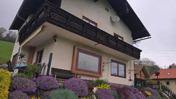 Traditionelles Haus mit Holzbalkon und blühendem Vorgarten vor einer malerischen Bergkulisse.