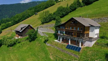 Luftaufnahme von zwei traditionellen Holzhäusern auf einem grünen Berghang mit weitem Blick auf die umliegende Landschaft.