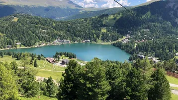 Malerischer Bergsee umgeben von bewaldeten Hängen und einem idyllischen Dorf unter blauem Himmel.