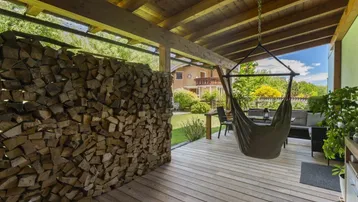 Gemütliche überdachte Holzterrasse mit Hängesessel und einem dekorativen Holzstapel als Sichtschutz.