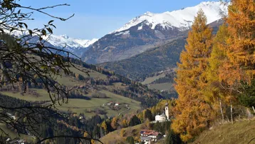Panoramablick auf schneebedeckte Berge und ein malerisches Tal mit Herbstbäumen und einer Kirche.