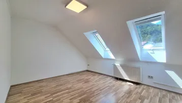 Helles Dachzimmer mit schrägen Wänden, zwei Dachfenstern und Holzboden, ideal als Schlaf- oder Arbeitszimmer.