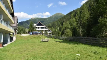 Mehrere Gebäude in einer alpinen Landschaft mit grünen Wiesen und bewaldeten Bergen im Hintergrund.