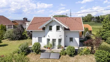 Weißes Einfamilienhaus mit rotem Ziegeldach und Solarpaneelen auf einem grünen Grundstück.
