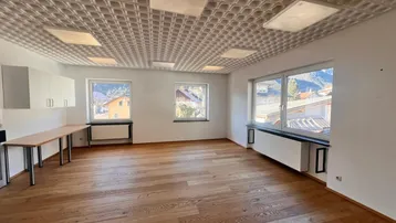 Geräumiges Büro mit Holzboden, großen Fenstern und strukturierten Deckenplatten, ideal für Arbeitsbereiche.