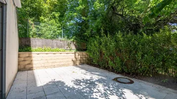 Sonniger Terrassenbereich mit Steinplatten, umgeben von üppigem Grün und einer Natursteinmauer.